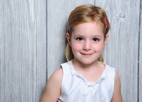 Porträtt av en söt fyra år leende blond flicka i vit klänning och färgglada barrettes i hennes hår står på papper grå trästaket bakgrund — Stockfoto