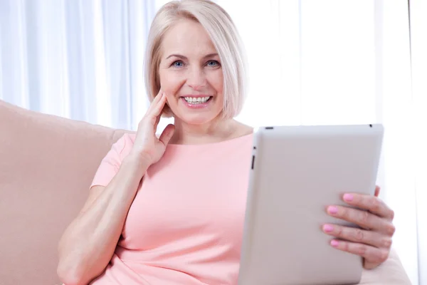 Uśmiechający się piękna kobieta w średnim wieku siedzi na kanapie z tabl — Zdjęcie stockowe
