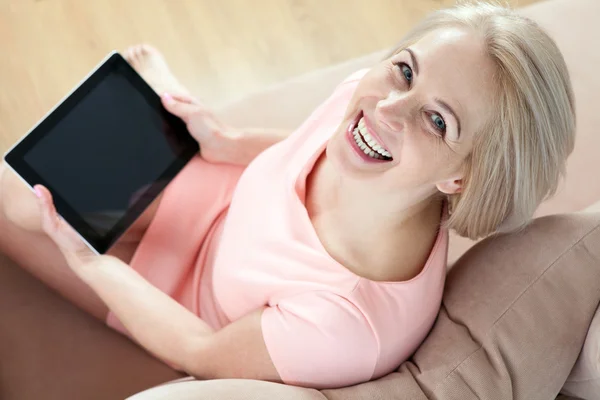 Smiling beautiful middle-aged woman sitting on couch with a tabl — Stock Photo, Image
