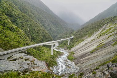 Otira Viaduct Gözcü Arthurs Geçidi, yeşil dağlar arasında engebeli bir vadiyi geçer, aşağıdan akan bir nehir ve uzaktaki sisli tepeler.