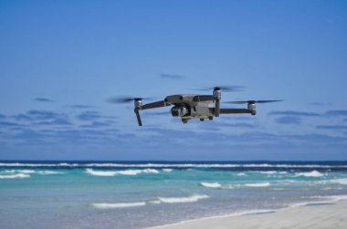 Bir insansız hava aracı turkuaz bir okyanus kıyısı üzerinde süzülür, açık güneşli bir günde dalgaların ve kumlu sahillerin hava görüntülerini yakalar, seyahat, teknoloji ve açık havada keşif çağrıştırır..