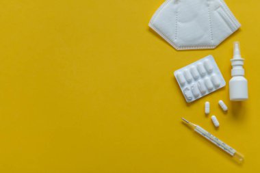 FFP3 maskesi, burun damlası, termometre, tabletler, çeşitli ilaçlar masanın üzerinde duruyor, üst görünümde, sarı arka planda, düz yaylı.