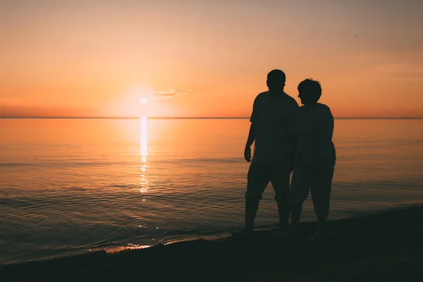 Silueta de pareja adulta camina en la orilla del mar contra una puesta de sol . — Foto de Stock