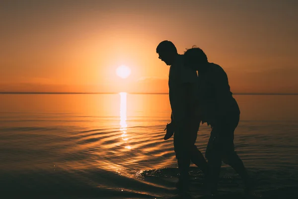 Silueta de pareja adulta camina en la orilla del mar contra una puesta de sol . — Foto de Stock