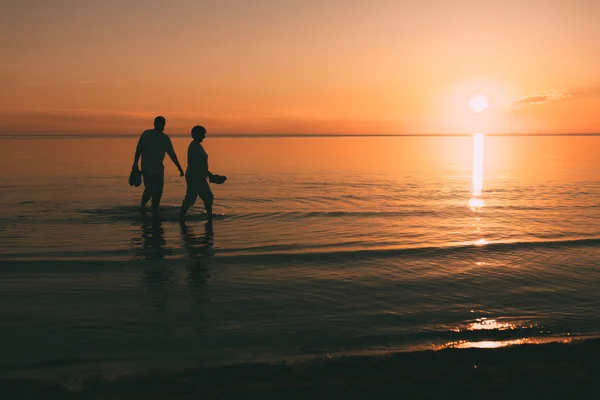 Silhouette eines erwachsenen Paares, das im Meer lebt und Schuhe in der Hand hält. — Stockfoto