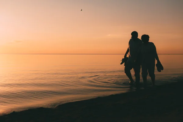 Silhouet van volwassen paar wandelingen aan de kust tegen een zonsondergang. — Stockfoto