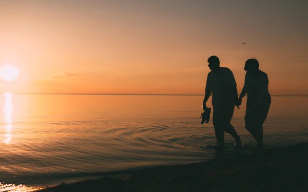 Silueta de pareja adulta camina en la orilla del mar contra una puesta de sol . — Foto de Stock