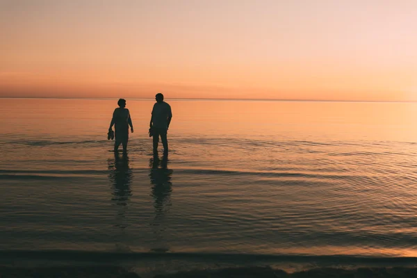 Силуэт взрослой пары, которая стоит в море и держит в руках обувь . — стоковое фото