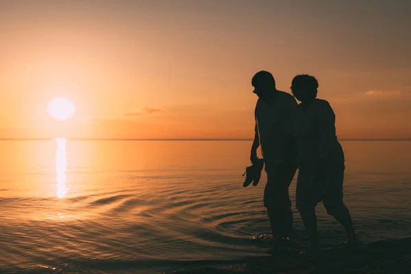 Silueta de pareja adulta camina en la orilla del mar contra una puesta de sol . — Foto de Stock