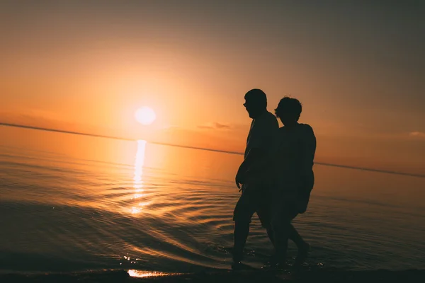 Silueta de pareja adulta camina en la orilla del mar contra una puesta de sol . — Foto de Stock
