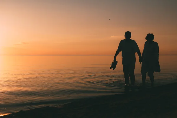 Silhouette of adult couple walks on the seashore against a sunset. — Stock Photo, Image