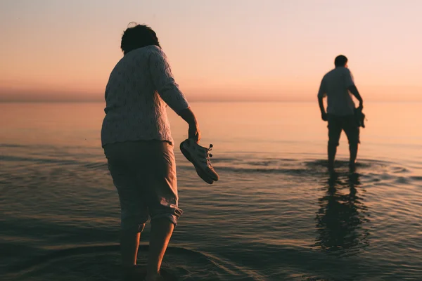 Małżeństwo dla dorosłych w ręku trzyma buty i spacery po wodzie. — Zdjęcie stockowe