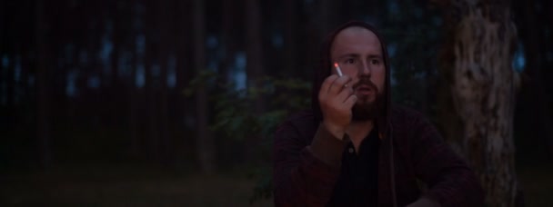 El hombre barbudo fuma cerca de una fogata en la naturaleza — Vídeos de Stock