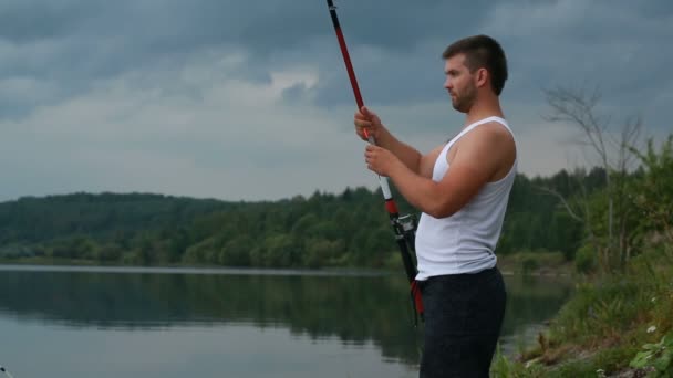 Jovem pescador com uma vara de pesca na margem do lago à noite — Vídeo de Stock