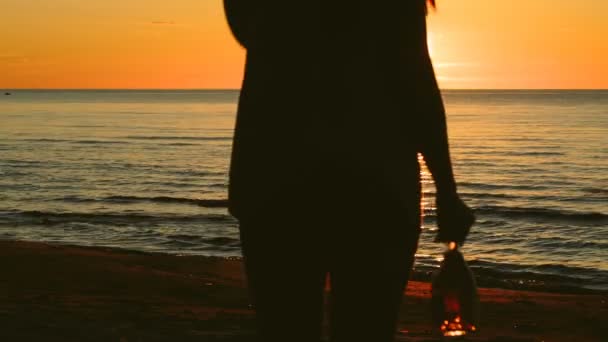 Ragazza con una bottiglia di vino in mano cammina lungo la spiaggia al tramonto — Video Stock