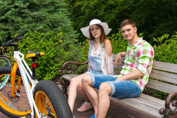 Jovem casal elegante sentado no banco perto de bicicletas no parque — Fotografia de Stock