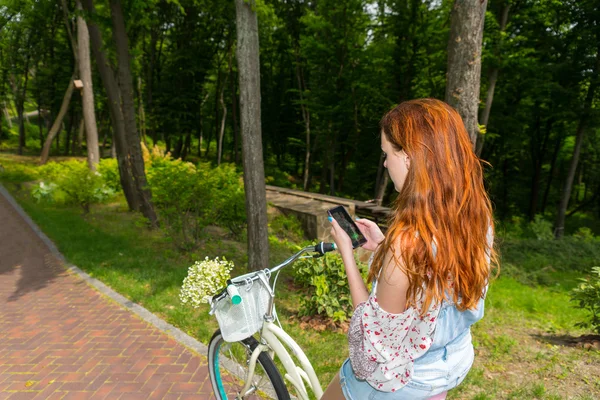 Jovem mulher sentada em sua bicicleta e usando seu telefone em um parque — Fotografia de Stock