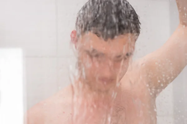 Chico tomando una ducha y sosteniendo la cabeza de la ducha detrás de vidrio nebulizado — Foto de Stock