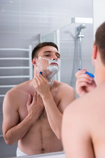 Homem fazendo a barba com um sorveteiro e olhando para o espelho no bathro — Fotografia de Stock