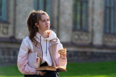 Düşünceli genç bir kadın, şehir parkında bir ekmeği abur cubur olarak yerken, kolunun altında ek kitaplarla oturuyor ya da ders çalışıyor.