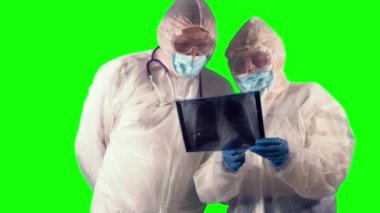 PPE üniformalı ve tıbbi koruyucu maskeli iki doktor yeşil arka plan üzerinde test yapmak için hastalarının röntgen filmlerini inceliyor.