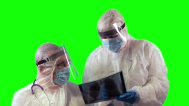 Tıbbi koruyucu maskeler ve yüz kalkanları takan PPE üniformalı iki doktor Covid üzerinde test yapmak ve yeşil arka plan üzerine notlar almak için hastaların röntgen filmlerini inceliyor.