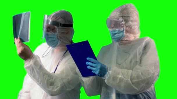 Dos médicos en uniformes de traje PPE, con máscara de protección médica y protector facial están inspeccionando una radiografía de los pulmones de los pacientes como una prueba en Covid y tomando notas sobre el fondo verde — Vídeos de Stock