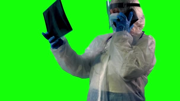 Læge eller sygeplejerske iført en PPE-dragt, ansigtsskærm og maske inspicerer en patienter røntgen af lunger som test på Covid, mens de taler på en telefon, på grøn baggrund – Stock-video
