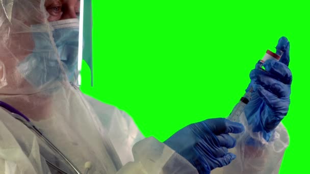 Врач в костюме СИЗ держит шприц и вакцину COVID-19 в голубых нитрильных перчатках, готовясь сделать инъекцию, на зеленом фоне — стоковое видео