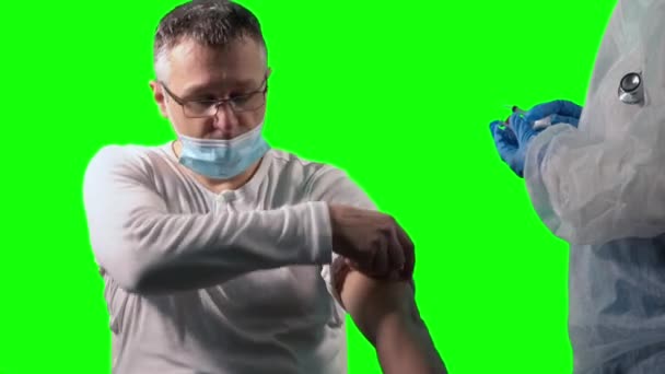 Врач в защитном костюме и защитном щите держит шприц и вакцину COVID-19 в голубых нитрильных перчатках, готовясь сделать инъекцию пациенту-мужчине, на зеленом фоне — стоковое видео