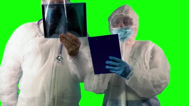 Лікар у формі костюма PPE перевіряє пацієнта на рентгенівському знімку легенів як тест на Ковиді, а інший робить нотатки на зеленому фоні. — стокове відео