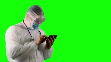 PPE üniformalı bir doktor, yüz kalkanı, maske ve steteskop elinde bir telefon tutuyor ve yeşil arka planda Covid ile ilgili haberleri arıyor.