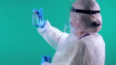 PPE giysisi ve yüz maskesi takan bir doktor, mavi arka planda Coronavirus tedavisi aşısı olan bir şişeyi sallıyor ve inceliyor.