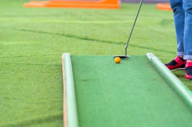 Yapay yeşil çimenlikte minyatür golf oynayan kişi bacaklarının, topun ve golf sopasının düşük açılı bir açıyla...