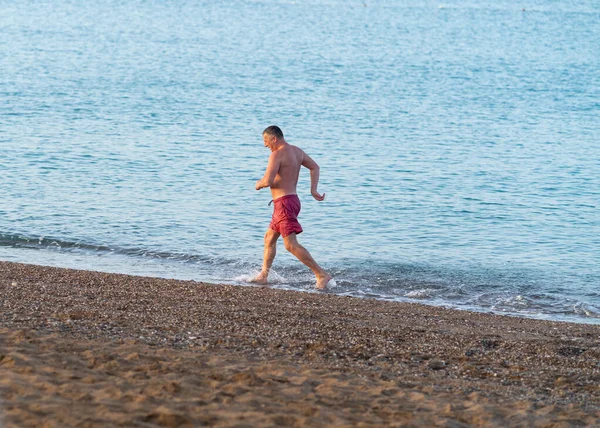 健康的なアウトドアライフスタイルのコンセプトで彼の水着で海の端に浅い波のビーチに沿ってジョギングアスレチック中年の男性 — ストック写真