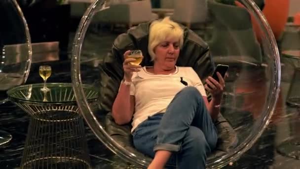 Жінка середнього віку розслабляється в готельному лаунжі зі своїм мобільним телефоном — стокове відео