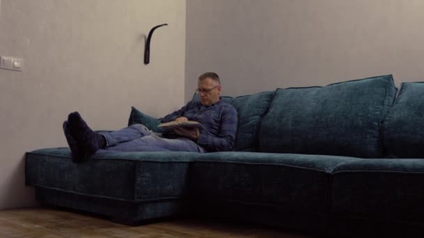 Hombre de mediana edad relajándose leyendo un libro por la noche en un sofá — Vídeos de Stock