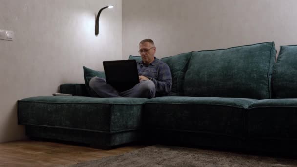 Orta yaşlı bir adam büyük bir kanepede dizüstü bilgisayarıyla dinleniyor. — Stok video
