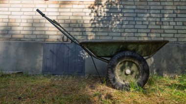 Lastik tekerlekler üzerinde metal bir bahçe arabası bir tuğla evin yanında boş duruyor..