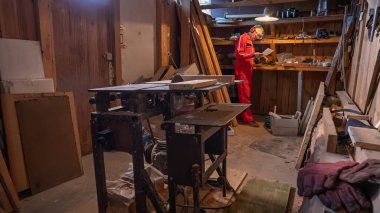 Bir atölyede, bir adam aletlerle rafların yanında dururken bir çizime ya da nota bakar..