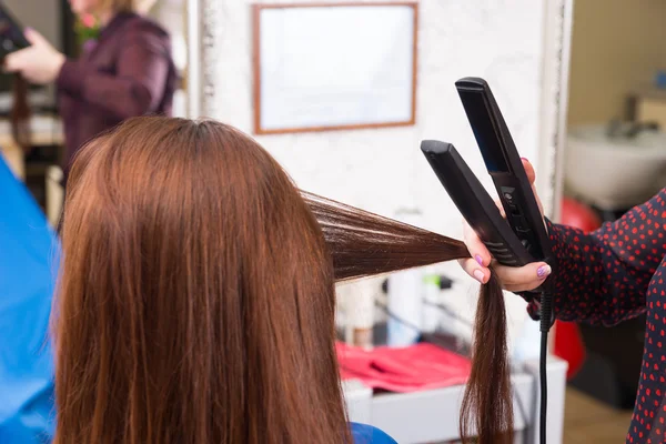 Estilista usando plancha plana en el cabello de cliente morena — Foto de Stock