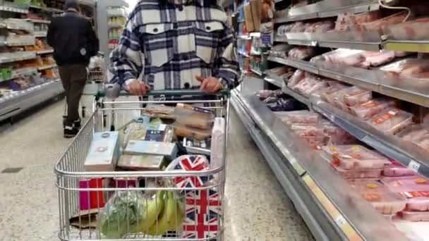 マンチェスター イギリス Circa 2021年3月 スーパーマーケットの顔のマスクを身に着けている人々の中で タータンジャケットの女性は生の肉棚でショッピングカートやトロリーを押している 彼女の後ろの店のアシスタントは注文を選んでいる — ストック動画