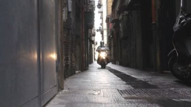 Yüksek binalar arasında karanlık dar bir ara sokak olan İtalyan şehri. Motosikletin düşük açılı görüntüsü ve sabit kameraya doğru gelen farlar. Plastik kalkanın arkasında tanınmayan bir bisiklet sürücüsü var..