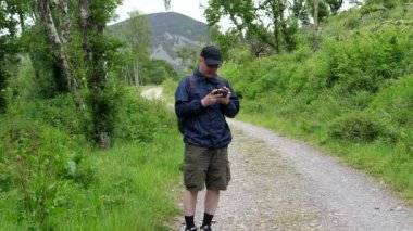 Sırt çantalı, gözlüklü bir adam dağdaki toprak yolda durup telefonunu kullanıyor. Muhtemelen harita uygulamasını ve el kamerasını kontrol ediyordur..