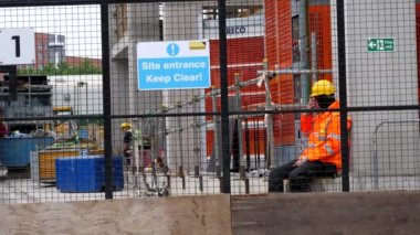 MANCHESTER, İngiltere, BİRLİK KINGDOM - CIRCA EPTEMBER, 2021: Güvenlik giysileri giyen trafik müdürü veya kapıcı inşaat veya inşaat alanının giriş kapısında çitin arkasında oturuyor.