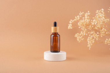 Beyaz podyumda bej arka planda kuru bitkilerle sunum yapmak için bambu kapaklı kehribar cam damlası şişesi..