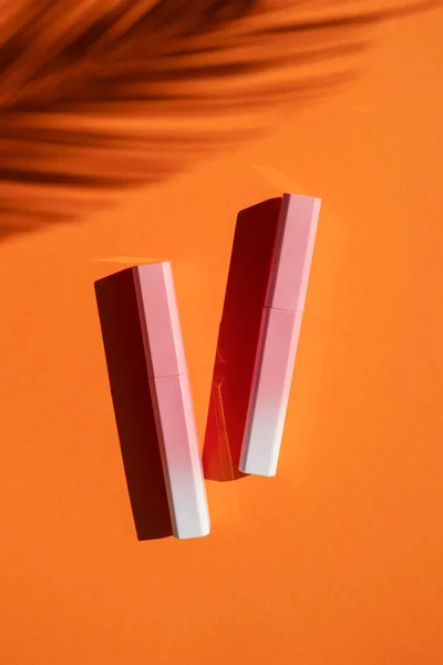 Lip gloss in a pink-to-white gradient case with palm leaves shadow. Top view on orange background. — Stock Photo, Image