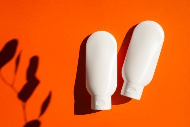 Turuncu arka planda krem ya da losyon için iki plastik beyaz tüp. Okaliptüslü üst manzara gölge bırakır. Cilt kremi kozmetik. Yüz bakımı için güzellik konsepti
