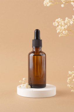 Beyaz podyumun üzerinde siyah kapaklı kehribar cam damlası şişesi bej arka planda kuru bitkilerle ürün sunumu için.