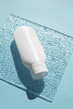 Mavi arka plan ve kıvrımlı cam üzerinde krem veya losyon için plastik beyaz tüp.